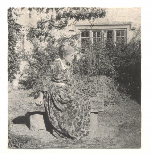 May Morris sits on a stone bench in a patterned dress in front of Kelmscott Manor.