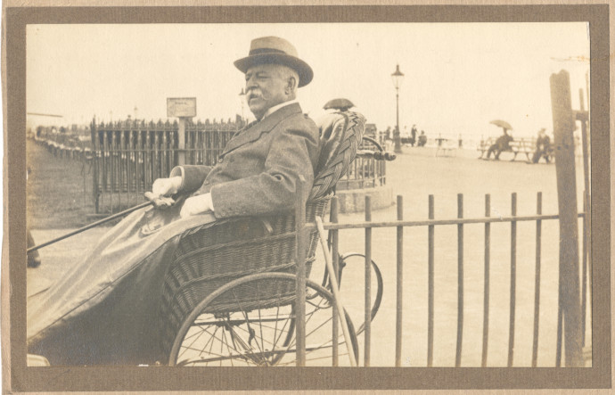 An elderly man sits in a wicker bath chair